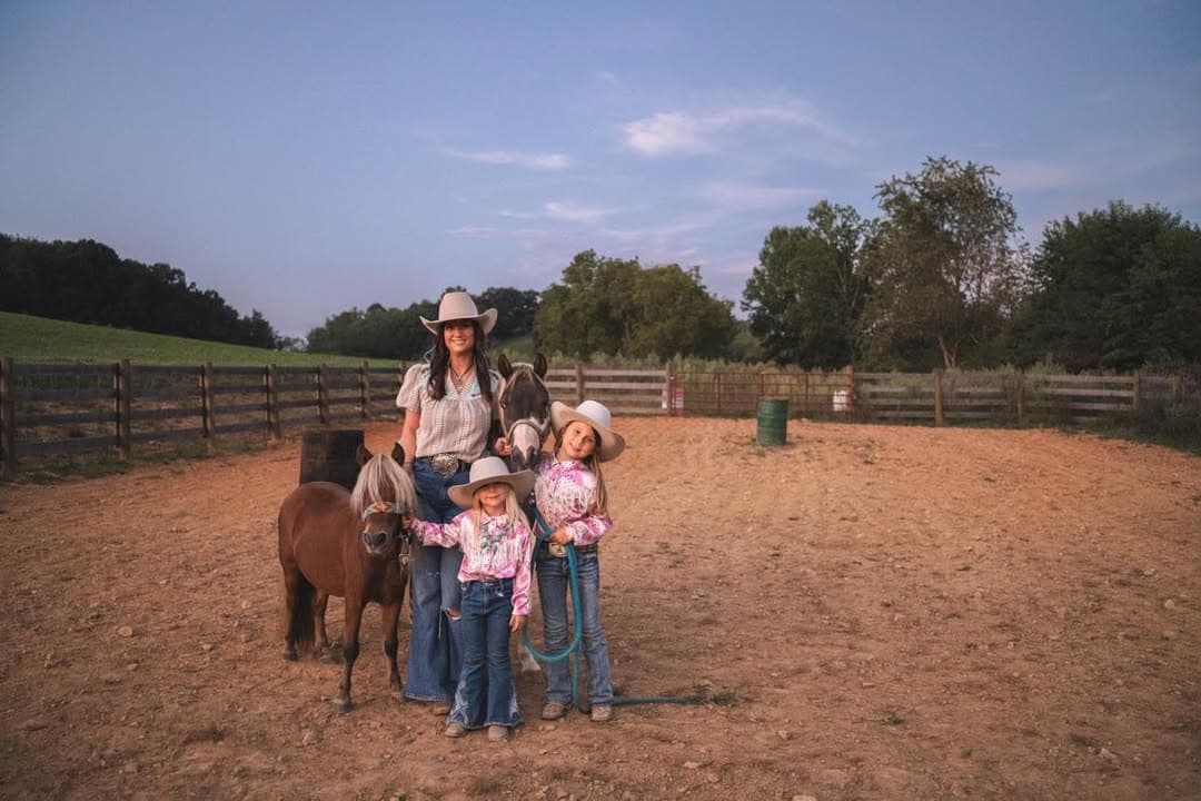 A mother and her two daughters in hats and show shirts standing with their pony and horse in the arena at golden hour