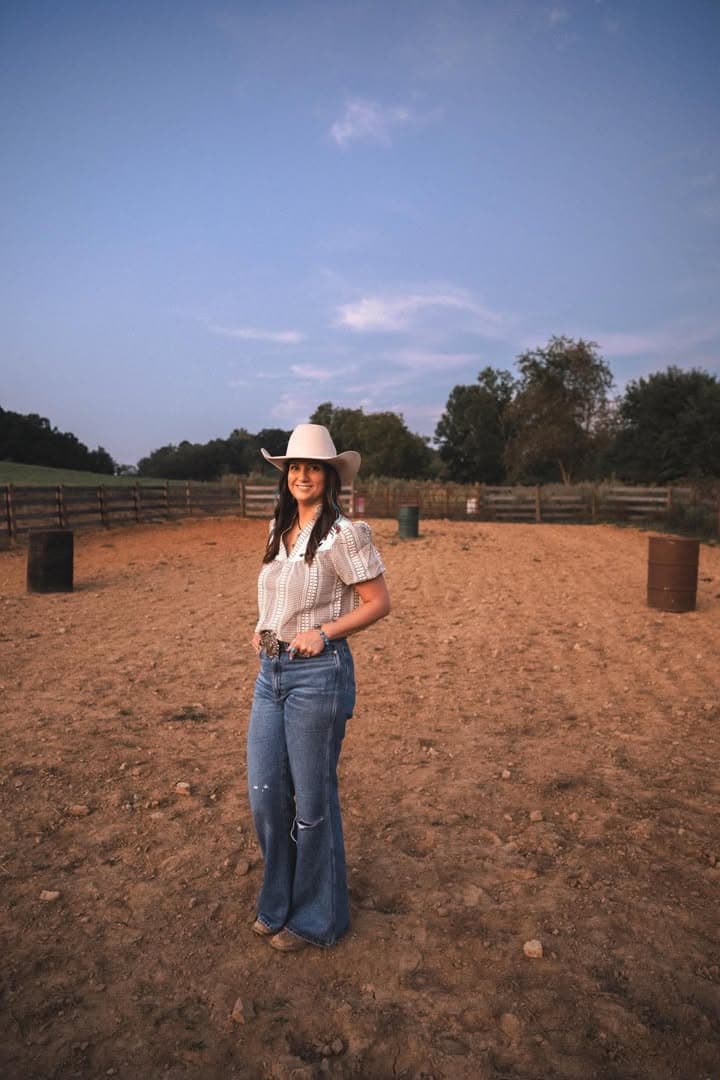 Clarissa Gosnell in a felt hat, standing in the arena at dusk with the barrels set behind her