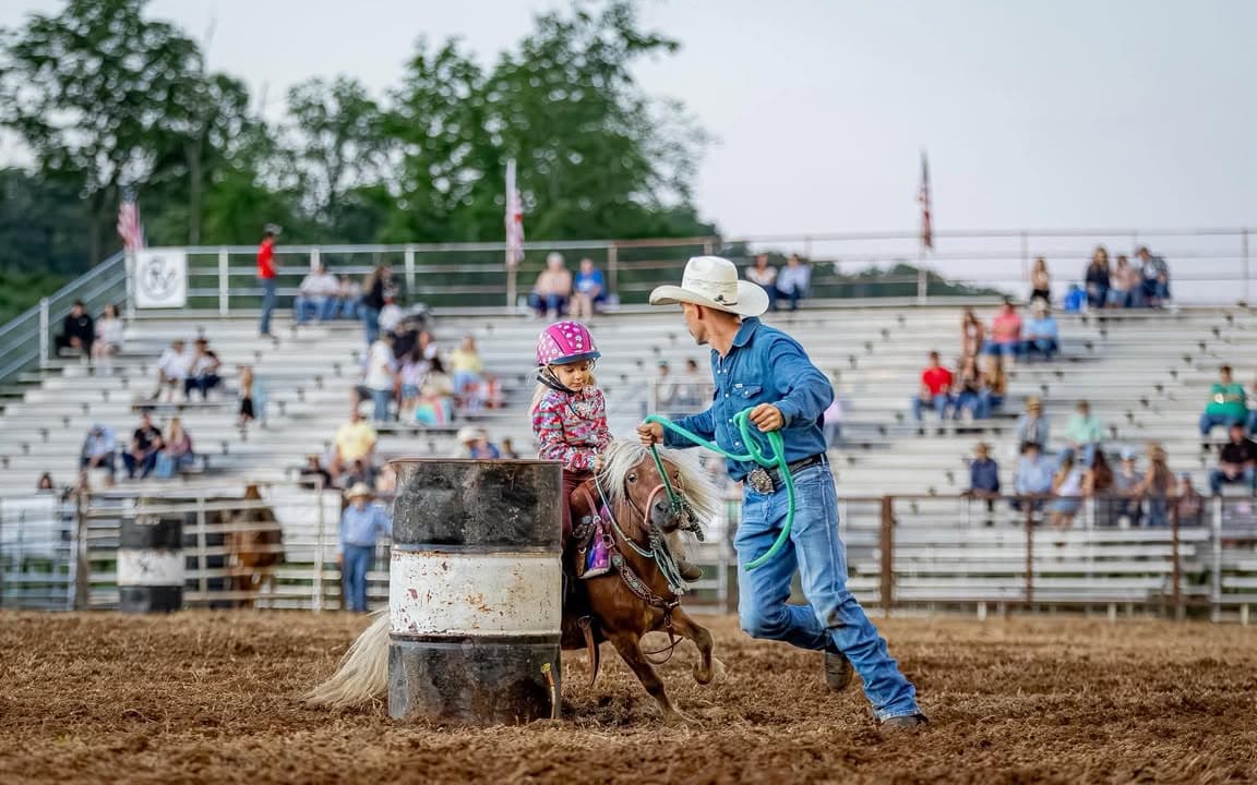 A lead line rider on a small pony rounding the barrel, led at a run by a helper in a cowboy hat, with the bleachers full behind them