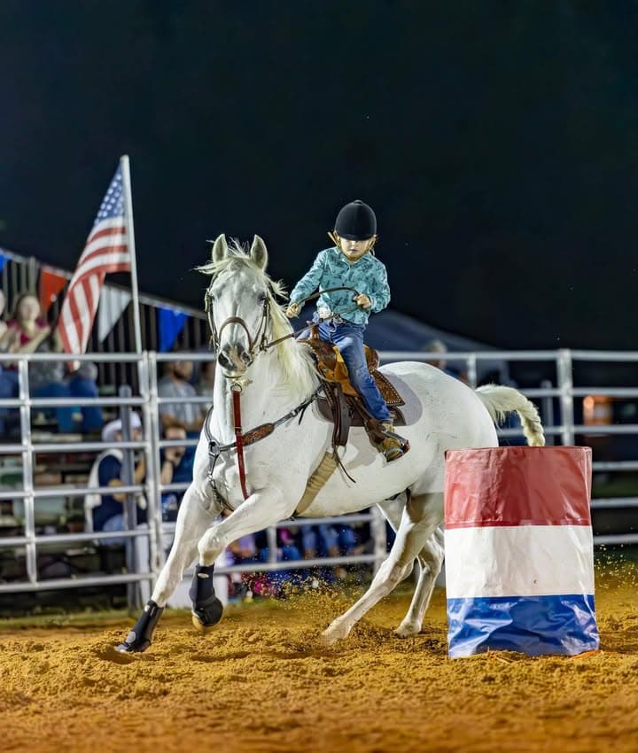 A young rider turning her grey horse around a red, white and blue barrel under the lights