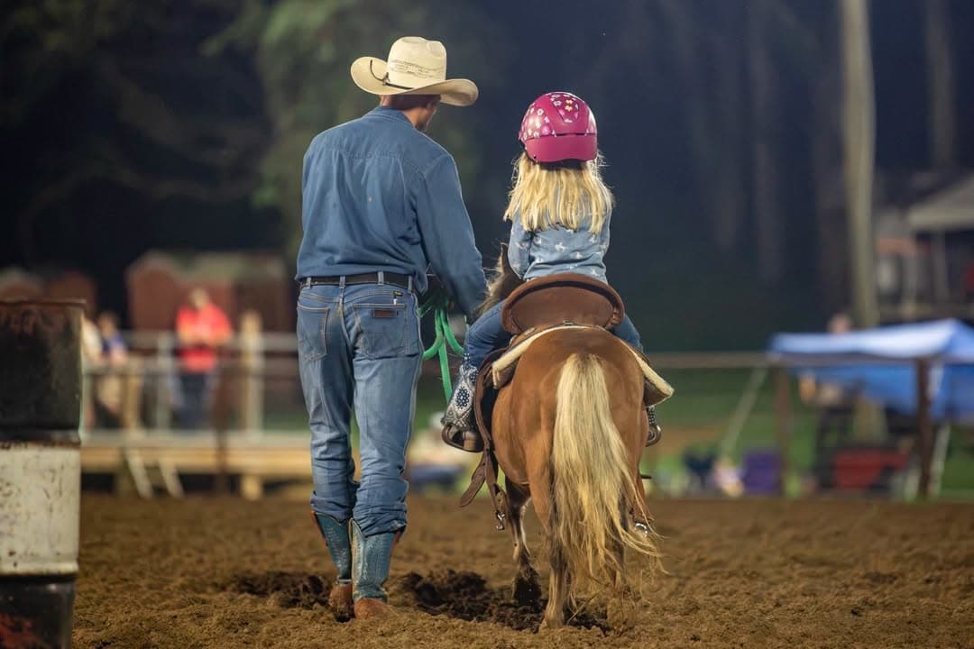 A helper walking beside a small rider on her pony as they head out of the arena at dusk